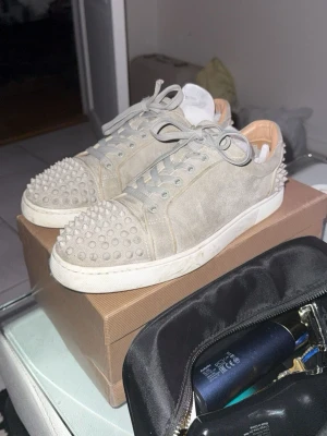 ÄKTA Grå Christian Louboutin sneakers med nitar - KVITTO FINNS! Säljer nu ett par ÄKTA gråa sneakers från Christian Louboutin med ikoniska röda sulor och coola nitar på tå och häl. Skorna är gjorda i mocka och har snörning samt rund tå. Perfekt för dig som vill sticka ut med en lyxig och edgy stil.