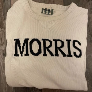 Vit stickad tröja från Morris - Snygg vit stickad tröja från Morris med svart logotyp broderad över bröstet. Tröjan har rund hals, ribbade muddar och lång ärm. Perfekt för dig som gillar klassisk stil med tydlig branding.