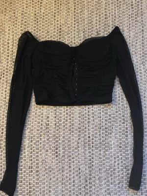 Svart långärmad korsett-topp - Säljer en svart korsett-topp med lång ärm och offshoulder-design. Toppen har snygg snörning framtill och är croppad med rynkade detaljer över bysten. Materialet känns mjukt och stretchigt. 