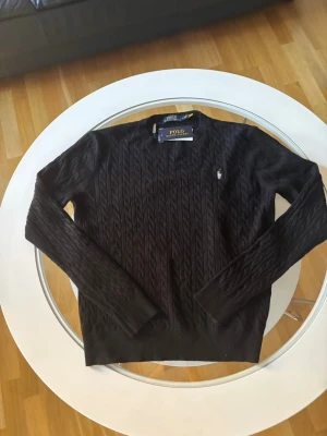 Svart kabelstickad tröja Polo Ralph Lauren helt ny - Svart kabelstickad tröja från Polo Ralph Lauren i storlek M. Tröjan har rund hals, långärmade ribbade muddar och den klassiska broderade loggan på bröstet. Materialet är mjukt och stickat, perfekt för kyliga dagar.