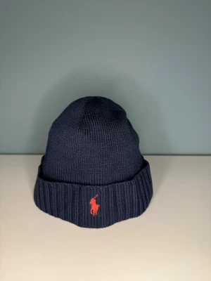 Mörkblå stickad mössa från Polo Ralph Lauren - Snygg mörkblå stickad mössa från Polo Ralph Lauren med klassisk röd logga broderad framtill. Mössan har bred uppvikt kant och är tillverkad i mjukt stickat material som håller dig varm. Perfekt för kalla dagar och enkel att matcha med din jacka. Sick 8/10. Vi kan gå ner i pris vid snabb och smidig affär. // Studio II Resell✅