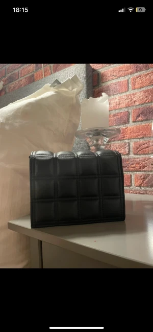 Svart quiltad handväska i skinn - Snygg svart handväska med quiltad design och rektangulär form. Väskan är tillverkad i skinn och har ett stilrent rutmönster på framsidan. Perfekt storlek för det viktigaste och passar till många olika looks. inget band till det