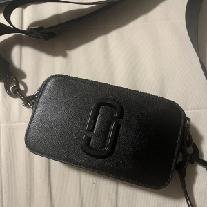 Svart axelväska från Marc Jacobs - Snygg svart axelväska från Marc Jacobs med en justerbar rem och dragkedja. Har haft väskan i 1 år. Dock har den gått sönder på dragkedjan som man kan se på 3e bilden, går att fixa hos butiker som har väskan inne. Har även en helt ny dragkedja som också visas på 3e bilden annars är väskan som ny. säljer den för 2200 just då för dragkedjan.