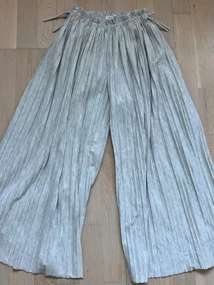Grå luftuga plisserade wide byxor från Zara - Säljer ett par ljusgrå slöna och luftiga fladdriga plisserade byxor från Zara för barn från ca 10-12. Byxorna har snörning i midjan och är tillverkade i ett mjukt bomullsmaterial med söta rosetter på sidorna. Perfekta för en chill och avslappnad stil och sköna för barn att ha ute på sommaren och våren. Har två små hål på ändan av en av benen som knappt märks, särskilt eftersom att de är så fladdriga. Pris kan diskuteras.