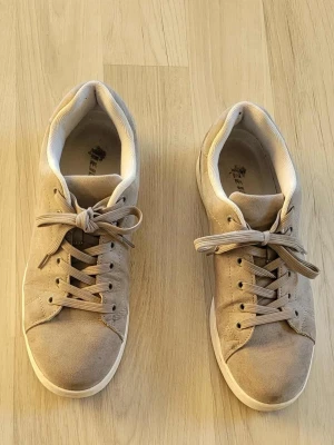 Beige sneakers i mocka med snörning - Snygga beige sneakers med rund tå och platt sula. Skorna har ett stilrent utseende med snörning och är tillverkade i mjuk mocka. Insidan är fodrad med ljus textil för extra komfort. Perfekta för dig som gillar en clean och enkel look.