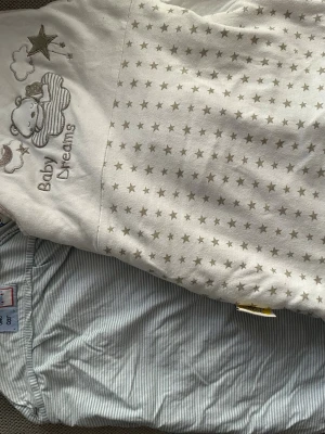 Vit babyväst med stjärnor och dragkedja - Säljer en söt vit babyväst från Baby Dreams med beige stjärnmönster och broderad nalle på framsidan. Västen har dragkedja och insida med färgglada små bilar och stjärnor. Utsidan är mjuk och tillverkad i bomull, perfekt för små barn.
