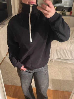 Svart sweatshirt med half zip - Svart sweatshirt med vit half zip-dragkedja och hög krage. Tröjan har en loose passform och är långärmad med ribbade muddar vid ärmslut och nederkant. Perfekt för chill dagar och enkel att styla med jeans.
