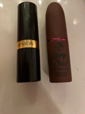 Revlon läppstift & W7 Lippy Chic - Två läpprodukter: ett klassiskt svart och guld läppstift från Revlon samt ett brunt, matt flytande läppstift från W7 Lippy Chic med rosa detalj. Perfekt för dig som gillar både traditionella och flytande läppfärger.