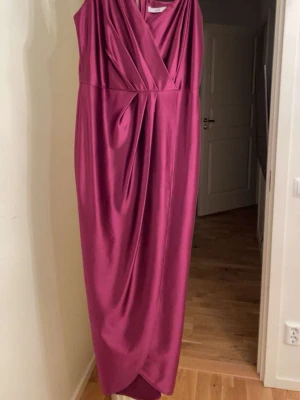 Lång vinröd klänning från WalG - Säljer en elegant vinröd långklänning från WalG med omlott-design och v-ringad hals. Klänningen är ärmlös och har ett snyggt fall i glansigt satinliknande material. Perfekt för dig som vill sticka ut med en stilren look.