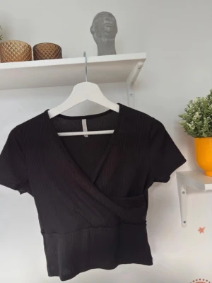 Svart omlott ribbad topp - Säljer en svart omlottopp från H&M med ribbad struktur och korta ärmar. Toppen har en snygg v-ringning och croppad passform som ger en modern look. Perfekt att matcha med jeans eller kjol för en cool stil.