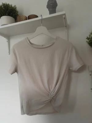 Beige ribbad topp med knyt - Säljer en beige ribbad topp med korta ärmar och snygg knytning framtill. Toppen har en rund halsringning och är gjord i mjukt material som känns skönt mot huden. Perfekt att matcha med jeans eller kjol för en chill look.