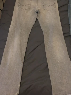 Grå jeans från Jack & Jones - Säljer ett par ljusgrå jeans från Jack & Jones Jeans Intelligence. Jeansen har raka ben, klassisk femficksdesign och subtila slitningar vid fickorna. Materialet är mjukt denim och färgen är snyggt tvättad för en avslappnad look. Finns litet hål över högra bakficka, annars är den i topp skick. Storlek 30/32