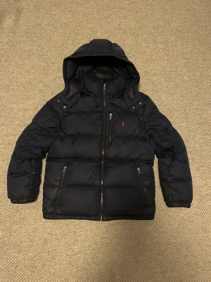  Kvitto finnsMörk blå pufferjacka från Polo Ralph Lauren - Säljer en svart pufferjacka från Polo Ralph Lauren med huva och flera dragkedjor. Jackan har det klassiska röda Polo-logot broderat på bröstet, praktiska fickor med dragkedja och snygga detaljer vid axlarna. Perfekt för kalla dagar och riktigt stilren.