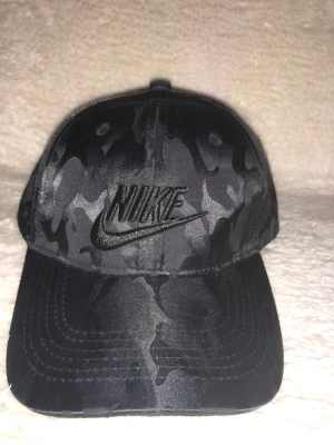 Svart keps från Nike - Snygg svart keps från Nike med diskret camouflage-mönster och broderad logga framtill. Justerbar rem baktill för perfekt passform. Perfekt för dig som vill ha en sportig och cool stil.
