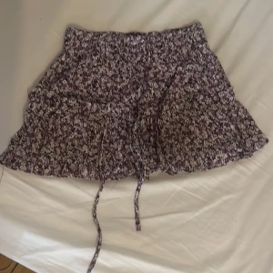 Ljuslila blommig volangkjol med snörning - Supersöt kort kjol med blommigt mönster i lila, vitt och svart, från Zara. Den går att knyta tajtare med snören framtill och är perfekt för en fin sommardag. Har använts en del men visar inga synliga tecken på skador eller annat. Säljer för 50 kr.