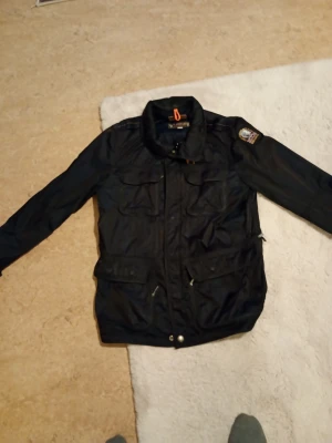 Design jacka Svart vindjacka Parajumpers XL - Svart vindjacka från Parajumpers i storlek XL. Jackan har flera fickor med dragkedja och knappar, samt en patch på ena ärmen. Materialet är syntet och jackan har meshfoder för ventilation. Perfekt för dig som gillar funktionell och snygg streetstil.