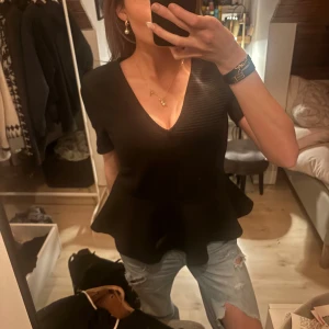 Svart peplum topp med v-ringning - Säljer en svart peplum topp med v-ringning och korta ärmar. Toppen har en markerad midja och volang nertill som ger en snygg siluett. Perfekt att styla med jeans eller kjol för en trendig look.
