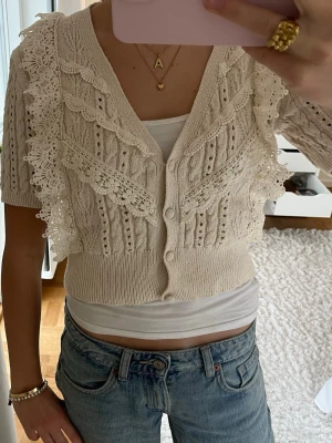 Beige stickad kofta från Zara - Superfin beige stickad kofta från Zara med korta ärmar och v-ringning. Koftan har dekorativa spetsdetaljer och knappar framtill. Den är croppad och har ett snyggt mönster i stickningen.