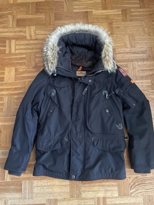Svart Parajumpers parkajacka med päls - Svart parkajacka från Parajumpers, modell Anchorage, med äkta päls på huvan och flera praktiska fickor. Jackan har broderad patch på ärmen, dragkedja och knappar framtill samt quiltad insida. Slim fit och perfekt för kalla vinterdagar.
