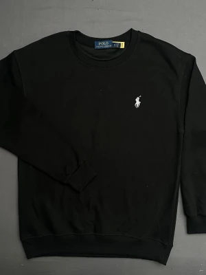 Svart sweatshirt från Polo Ralph Lauren - Svart sweatshirt från Polo Ralph Lauren med klassisk rund hals och broderad vit logga på bröstet. Tröjan har ribbade muddar vid ärmslut och nederkant, vilket ger en snygg och bekväm passform. Perfekt för dig som gillar stilrena och tidlösa plagg.