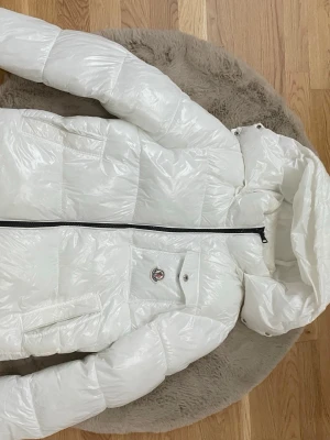 Vit Maya från Moncler - Säljer en vit pufferjacka från Moncler med glansig finish och huva. Jackan har en svart dragkedja framtill, praktiska fickor och Moncler-logga på bröstet. Perfekt för kalla dagar och riktigt snygg till streetwear.