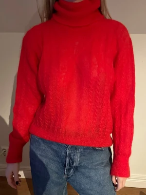 Röd polotröja i stickad ull - Säljer en snygg röd polotröja med stickad struktur och kabelmönster. Tröjan har lång ärm och hög polokrage som ger extra värme. Perfekt att matcha med jeans för en chill look. Materialet känns mjukt och är troligtvis ull.