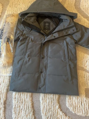 Canada Goose - Säljer en grå dunjacka från Canada Goose med huva och raka linjer. Jackan har både dragkedja och knappar framtill, samt två stora fickor. Medföljer en lös pälskant till huvan. Perfekt för kalla dagar och riktigt snygg stil.