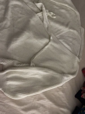 Vit omlott kofta med knytband - Säljer en vit stickad kofta med omlott-design och knytband i midjan. Koftan har långa ärmar och ribbade muddar, perfekt för lager-på-lager. Den mjuka stickningen gör den både skön och snygg att ha på sig.