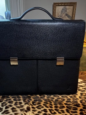 Louis Vuitton svart portfölj, väska, briefcase, herr, vintage - Säljer en klassisk svart portfölj från Louis Vuitton i strukturerat skinn. Väskan har två framfickor med silverfärgade spännen, ett rymligt huvudfack och flera innerfickor. Perfekt för laptop och dokument, med stilren design och diskret LV-logo i hörnet. 40 x 34 cm. Kan även tänkas byta!