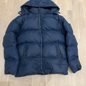 Mörkblå pufferjacka från Tommy Hilfiger - Snygg mörkblå pufferjacka från Tommy Hilfiger med huva och dragkedja framtill. Jackan har två fickor med dragkedja och klassisk logga på bröstet. Perfekt för kalla dagar och riktigt skön att bära.