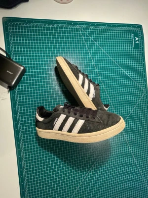 Adidas Campus sneakers i mörkgrå mocka - Säljer ett par Adidas Campus sneakers i mörkgrå mocka med klassiska vita ränder och vit häl. Skorna har svart snörning och en beige platt sula. Perfekta för dig som gillar retrostil och streetwear. Size 39,5