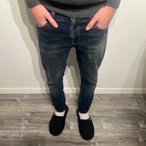  Nudie Jeans - Snygga mörkblå slim jeans från Nudie Jeans med klassiska orange sömmar och slitningar på framsidan. Modellen har fem fickor, normal passform i midjan och är tillverkad i mjukt bomullsmaterial. Perfekt för dig som gillar en smal siluett och stilrena detaljer. De har ett litet hål vid låret vilket enligt mig gör jeansen snyggare, sen är dragkedjan trasig men jag har sytt ihop den så de går att använda perfekt 👌 