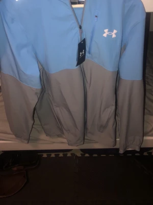 Grå och ljusblå vindjacka från Under Armour - •Säljer en snygg vindjacka från Under Armour i grått och ljusblått.  • Den är helt ny och är använd 0 gånger. • har prislappen och kvittot kvar där ni kan se på bilden. • Och det är storlek M. • Använd 0 gånger