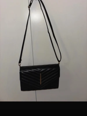 Svart quiltad axelväska med gulddetalj - Snygg svart axelväska i skinnimitation med quiltad framsida och guldfärgad metallstängning. Väskan har ett justerbart axelband men metallen som ska vara guldig har tappat färgen lite.