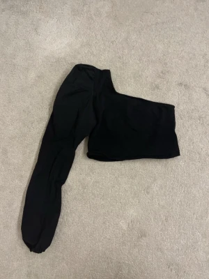 Svart oneshoulder topp  - Säljer en svart oneshoulder topp från Bershka i storlek S. Toppen har en lång ärm på ena sidan och ärmlös på andra, vilket ger en cool asymmetrisk look. Toppen är croppad 