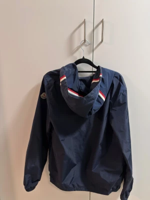 Mörkblå vindjacka från Moncler - Snygg mörkblå vindjacka från Moncler med huva och dragkedja. Jackan har Moncler-logga på ärmen och coola rödvita detaljer på huvan. Tillverkad i lätt polyester, perfekt för blåsiga dagar. Två fickor med tryckknappar och elastiska muddar vid ärmslut.