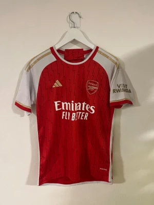 Arsenal 2023/24 hemma matchtröja  - Säljer en Arsenal matchtröja från adidas i storlek S. Tröjan är röd med vita och guldiga detaljer, har blixtmönster och klubbmärke på bröstet. Slim fit-modell med rund hals och korta ärmar. Materialet är lätt polyester, perfekt för fotboll.