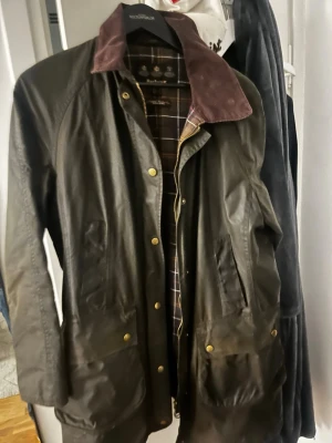 Barbour olivgrön parkajacka med rutig insida - Säljer en klassisk olivgrön parkajacka från Barbour med rutig insida och brun manchesterkrage. Jackan har stora fickor med tryckknappar och dragkedja framtill. Perfekt för dig som gillar tidlös stil och vill ha något som funkar året runt.