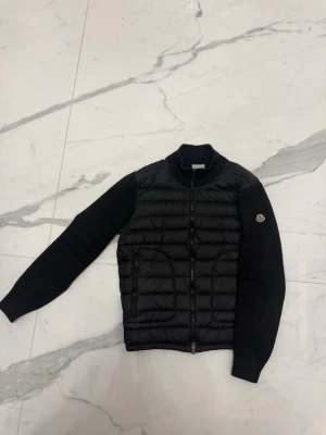 Svart cardigan Moncler - Svart cardigan från Moncler i storlek M med quiltad front och stickade ärmar. Jackan har dragkedja framtill, ribbad krage och Moncler-logga på ärmen. Materialmix av polyester och stickad ull ger en schysst kontrast och modern vibe. Kvitto finns digitalt.