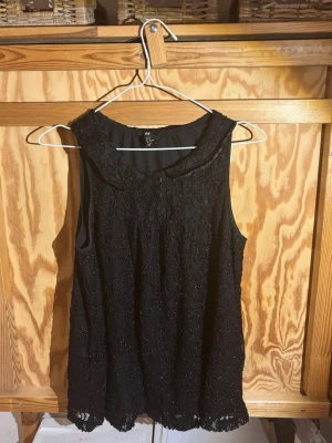 Svart glittrig spetsblus från H&M - Säljer en svart blus från H&M i storlek 42. Blusen är ärmlös och har ett glittrigt spetsmaterial med dekorativa knappar framtill. Rundad krage och lätt  spets tyg. Perfekt för dig som gillar detaljer och glitter.