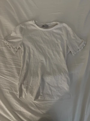 Vit t-shirt med pärldetaljer från Zara - Supersöt vit t-shirt från Zara i mjuk bomull. T-shirten har korta ärmar med unika pärldetaljer och spets längst ut på ärmsluten. Enkel rund halsringning och normal passform. Perfekt för dig som vill ha något basic men ändå extra. Pärlorna som är fästa vid ärmen har fått lite defekter vid användning samt tvätt som syns på sista bilderna. 