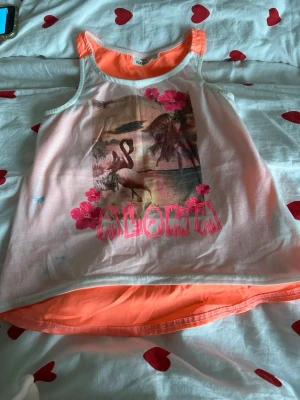 Aloha linnetopp från KappAhl - Säljer en färgglad linnetopp från KappAhl i storlek 134/140. Toppen har ett tropiskt tryck med flamingo, palmer och blommor samt texten 'ALOHA' i rosa. Framsidan är vit med tryck och baksidan är helorange. Perfekt för sommaren!