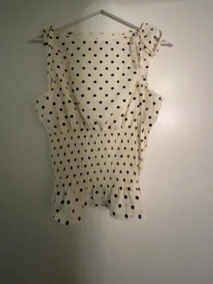 Prickig vit blus med rosett - Säljer en söt vit blus med svarta prickar och rosett på axeln. Blusen har ärmlös design, smockad midja och volang nedtill som ger en feminin touch. Perfekt för varma dagar och enkel att matcha med jeans eller kjol. Aldrig använd 