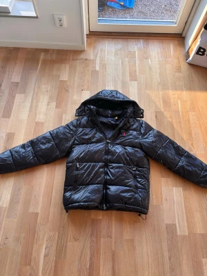 Svart pufferjacka från Polo Ralph Lauren - Svart pufferjacka från Polo Ralph Lauren med glansig finish och huva. Jackan har två framfickor med dragkedja och en röd Polo-logga på bröstet. Perfekt för kalla dagar och har en modern, sportig vibe. Fodrad och med quiltad design för extra värme.