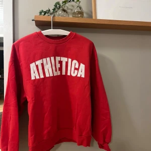 Röd sweatshirt ATHLETICA - Röd sweatshirt från ginatricot  med vit text framtill. Tröjan har rund hals, ribbade muddar vid ärmslut och nederkant samt lång ärm. Perfekt för dig som gillar sportig och avslappnad stil.