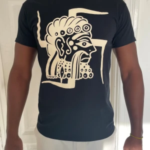 Svart t-shirt med grafiskt tryck Gildan - Svart t-shirt från Gildan i 100% bomull med ett stort grafiskt tryck i beige på framsidan som föreställer ett stiliserat ansikte och linjer. Klassisk rund hals och korta ärmar. Perfekt för dig som gillar unika motiv och streetwear.