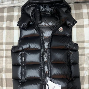 Svart dunväst från Moncler - Snygg svart dunväst från Moncler med glansig finish och klassisk logga på bröstet. Västen har hög krage, två dragkedjefickor och quiltad design. Perfekt för lager-på-lager och streetstyle. Stängs med dragkedja framtill.