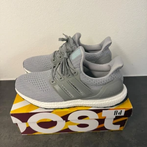 Adidas Ultraboost grå sneakers - Snygga Adidas Ultraboost sneakers i ljusgrå färg med stickad ovandel och klassiska tre ränder på sidan. Skorna har snörning, platt sula och en bekväm boost-mellansula i vitt. Perfekta för dig som gillar stilren och sportig look.