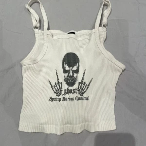Vit ribbad linnetopp med dödskalle - Vit ribbad linnetopp med smala axelband och tryck framtill av en dödskalle och skelett-händer. Texten 'ghost Spring Racing Carnival' under motivet. Croppad modell som passar perfekt till jeans eller kjol.