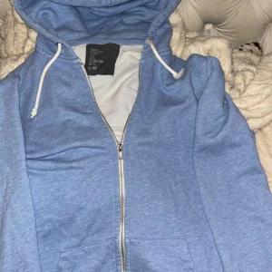 Ljusblå hoodie med dragkedja H&M - Ljusblå hoodie från H&M med hel dragkedja framtill och vit snörning i huvan. Tröjan har långa ärmar, fickor på magen och är gjord i mjuk bomullsmix. Perfekt för chill dagar eller när du vill vara extra comfy.